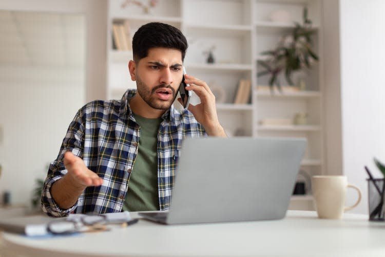 Confused man at his home desk talking on a mobile phone with his internet provider while sitting in front of his open laptop.