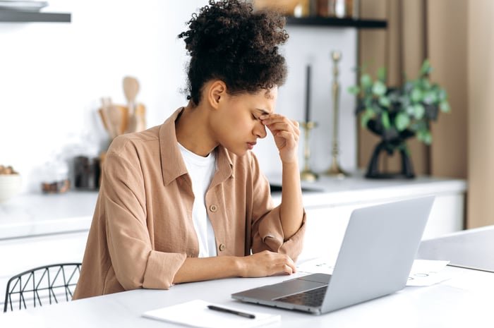 A person pinches the bridge of their nose while sitting at a table in-front of a laptop computer.