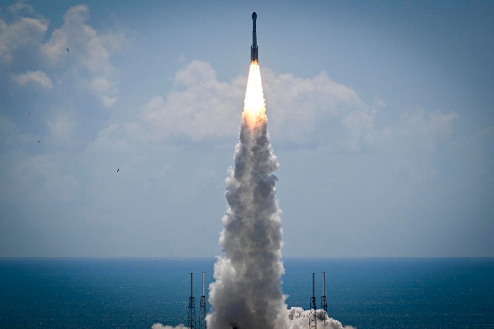 A rocket, seen from just above the launching pad, blasts skyward, with the ocean in the background and clouds scudding across the sky.