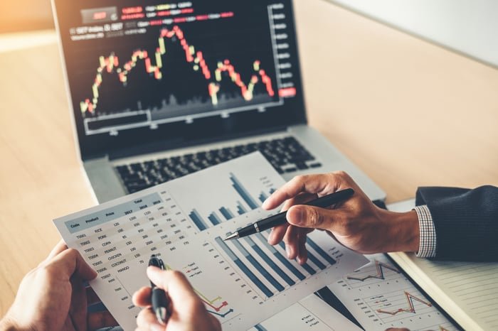 Two people analyzing business metrics on paper with a laptop in the background displaying a falling stock price chart.