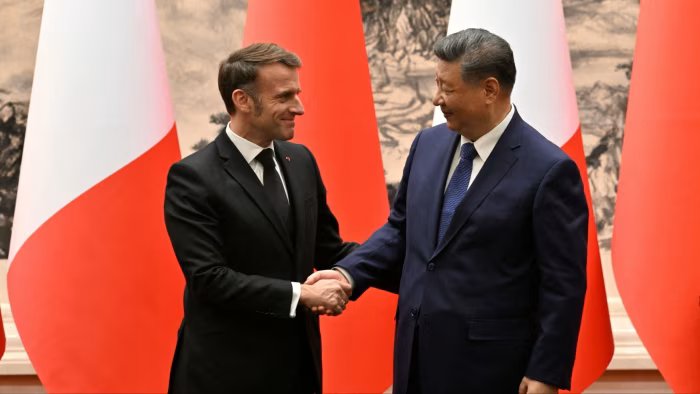 Macron and Xi shake hands and smile at each other in front of French and Chinese flags.