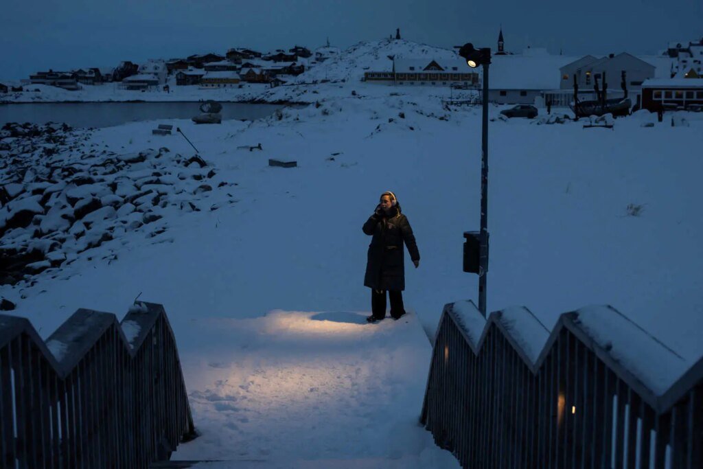 A person stands in the snow beneath a lantern, with a harbor in the distance and a snow-covered staircase in the foreground.