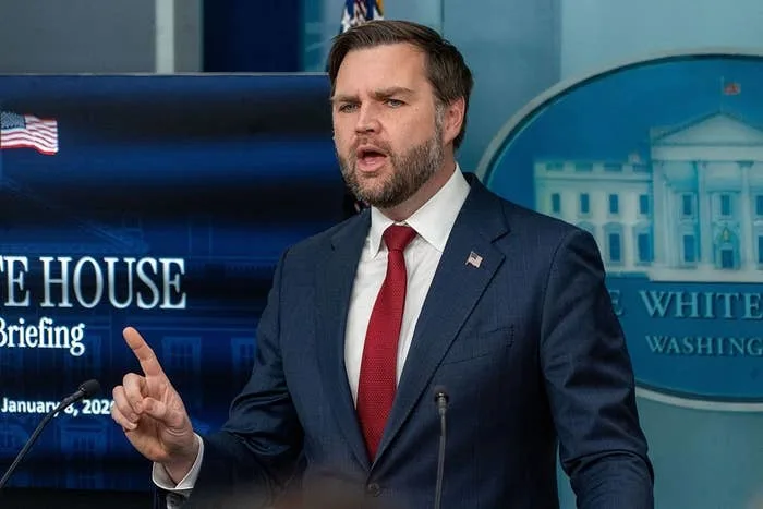 A person in a suit speaks at a White House press briefing, gesturing with one hand