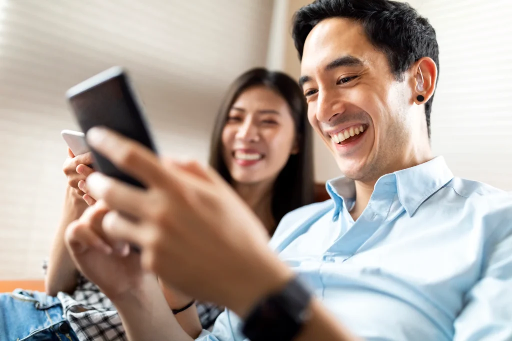 A couple is on a couch, smiling at their smartphone.