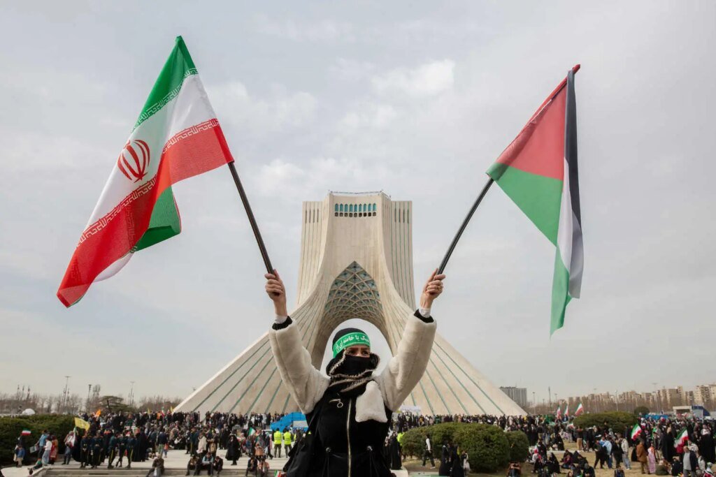 A person with a covered face and green headband with visible text raises two flags. A monument and large crowd are in the background.