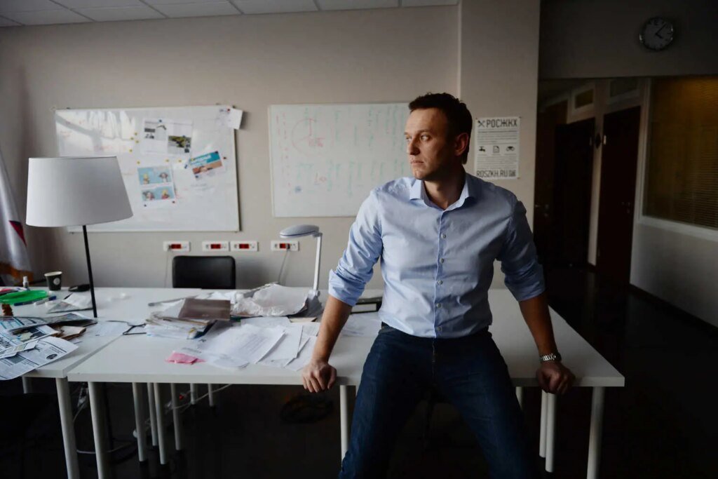 Aleksei A. Navalny sits on a white desk in a cluttered office. He wears a light blue shirt and dark pants, looking away with a neutral expression.