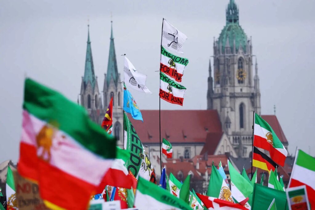 Numerous green, white and red flags, some reading "FREEDOM FOR IRAN," waving in front of two old stone church spires, on the left, and a domed building.