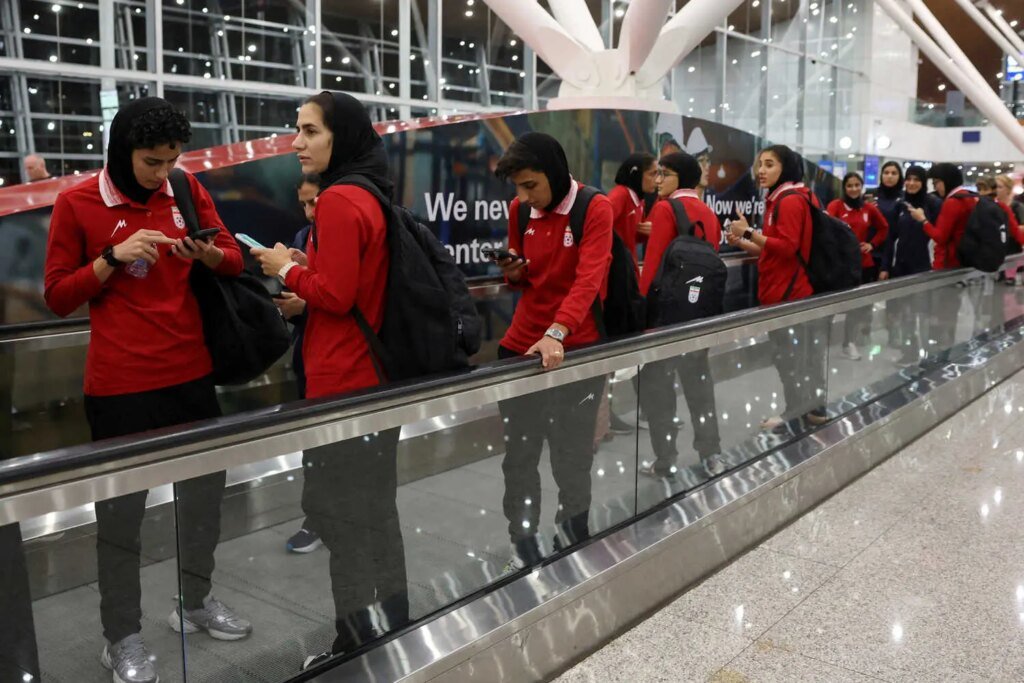 Women wearing red shirts and black head coverings stand on a moving walkway, some looking at phones.