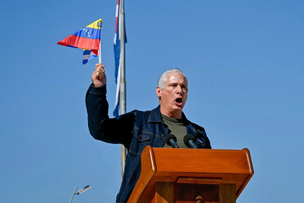 A person in a denim jacket stands at a podium, speaking with an open mouth. They raise a hand holding a small, colorful flag.