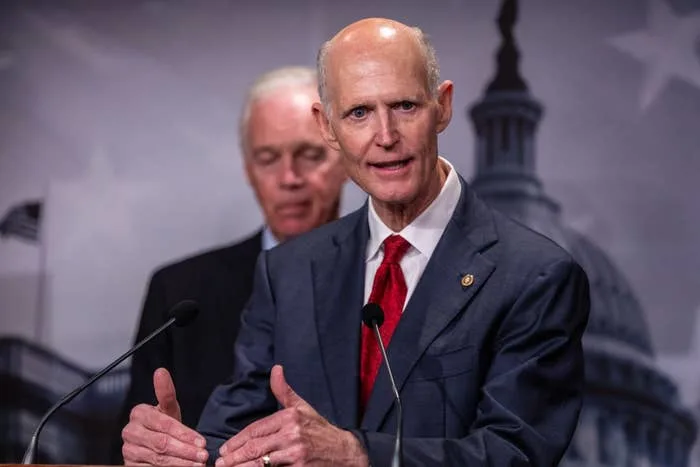 Two people standing at a podium, one speaking to an audience. Background shows a blurred Capitol building image