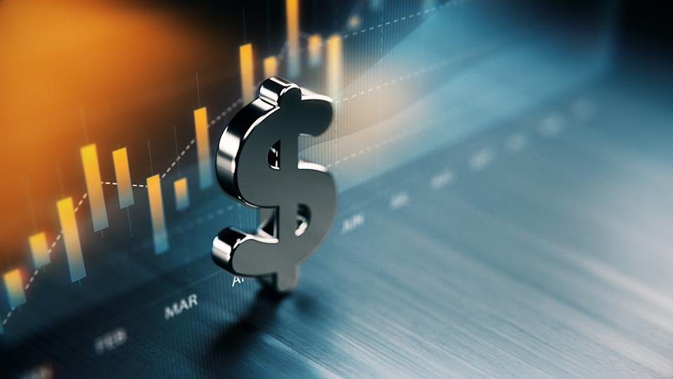 A photo illustration shows a silver 3-dimensional dollar sign sitting on a blue surface with a chart of bars in orange behind it.
