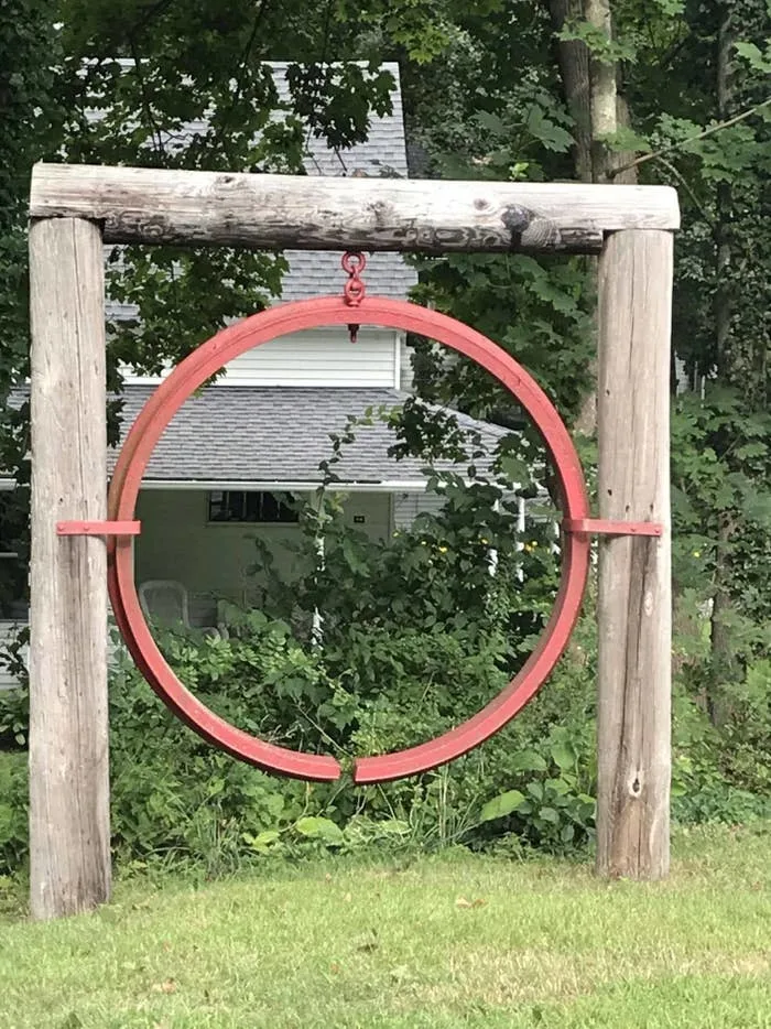 Wooden structure with a metal ring suspended in the center, set against a backdrop of trees and a house