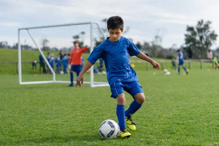 Young boy in soccer gear dribbles a ball on a field, with a goal and players in the background