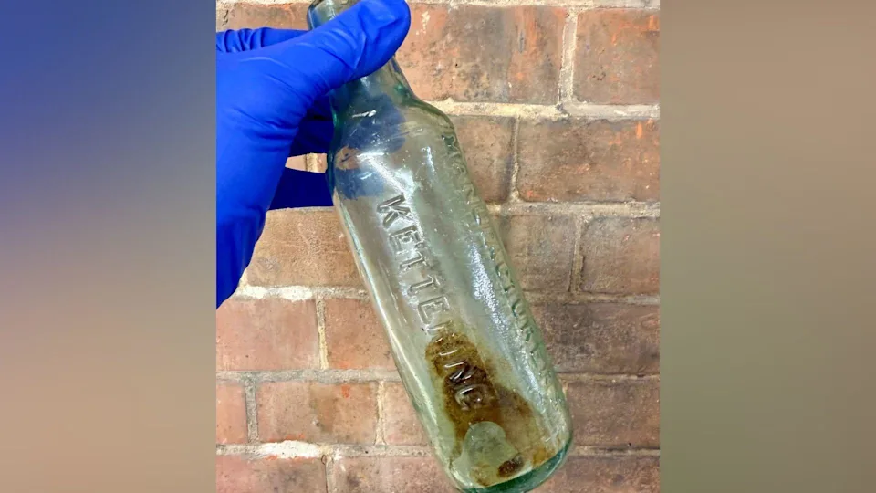An old empty glass bottle, being held up by a person wearing blue gloves. The bottle is being held up in front of a brick wall.