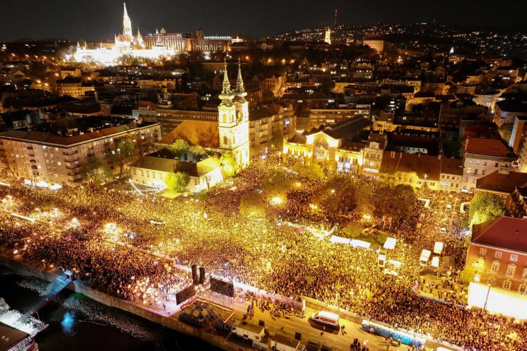 An aerial night view of a city showing streets densely packed with people. Bright yellow lights illuminate the crowd, with buildings and a river.