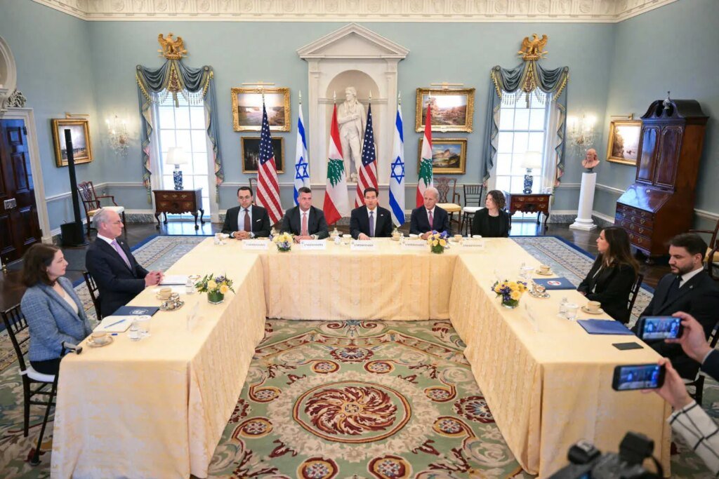 People are seated around a large U-shaped table in front of American, Israeli, and Lebanese flags.