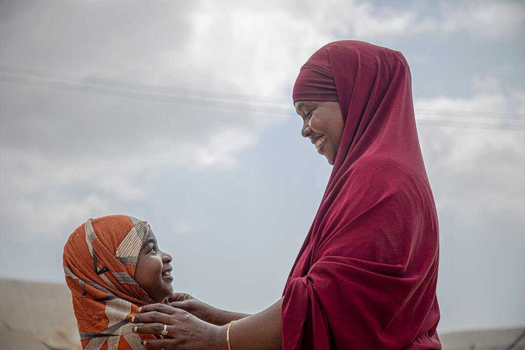 A woman in a red headscarf gently touches the head of a young girl wearing an orange headscarf, both smiling as they look at each other. The image symbolizes resilience and community resistance against female genital mutilation in Somalia.