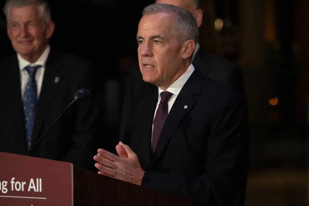 Mr. Carney, wearing in a dark suit, standing at a microphone. Another person in a dark suit smiles in the background.