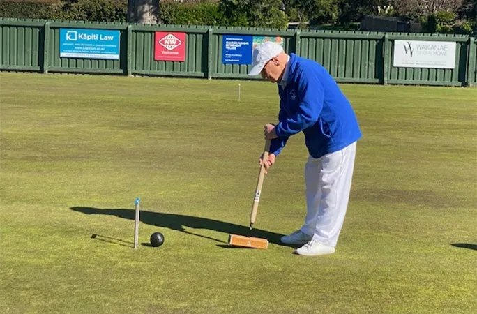 101-year-old Neville Sandiford in a white hat and blue jacket playing croquet on a green lawn.