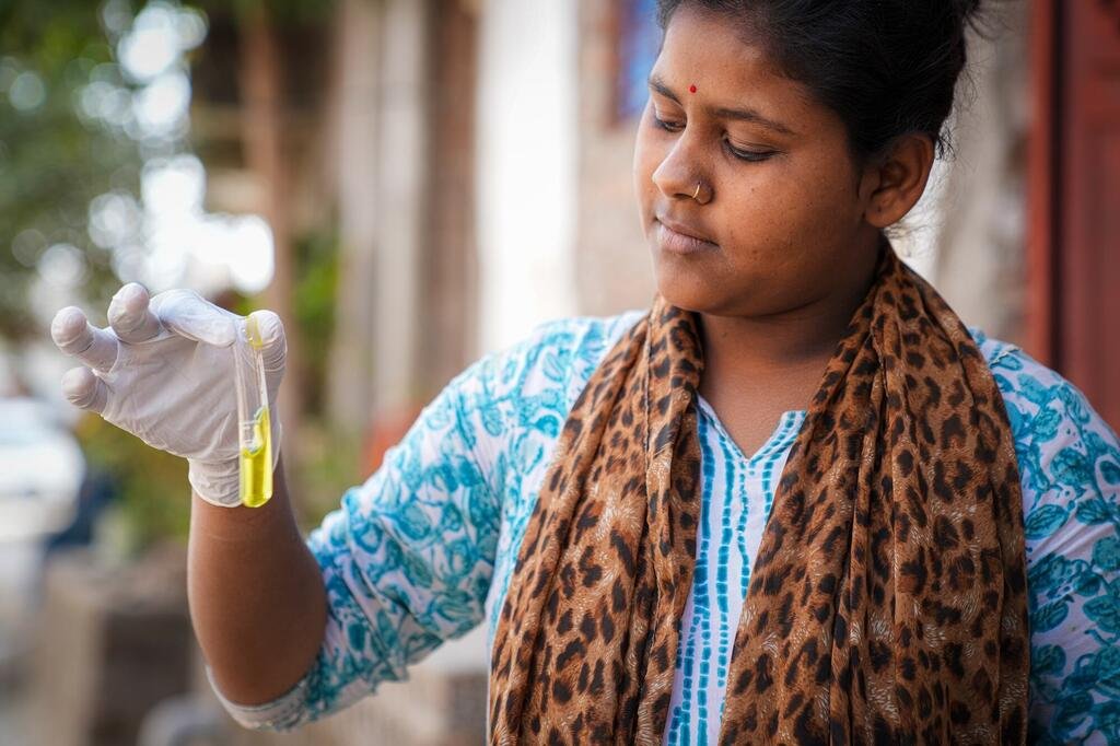 A fifteen-year-old Indian girl named Vandana carefully tests the quality of drinking water in her village, holding up a test tube with yellow liquid while wearing a white glove. She is part of the Jal Jeevan Mission, empowering communities to monitor water quality.