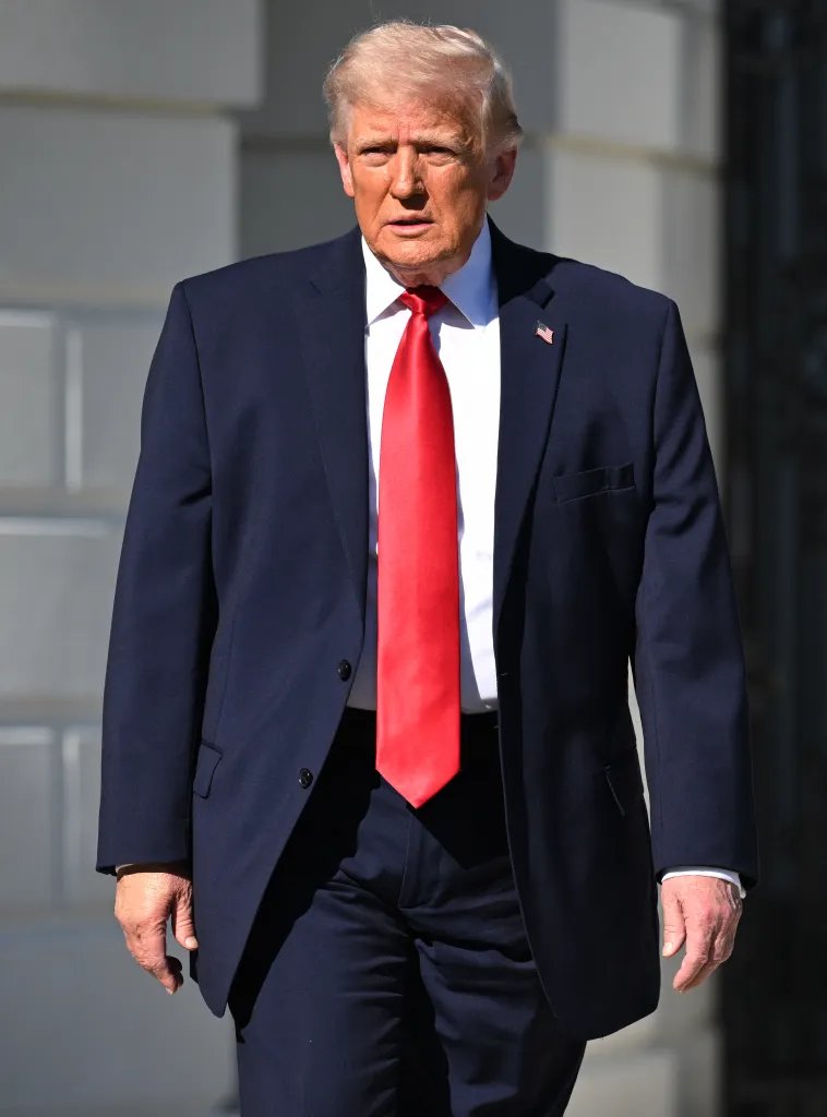 Donald Trump in a navy suit and red tie walking outdoors.